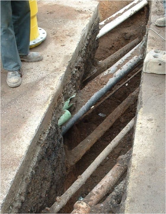 A trial hole in a footpath with multiple buried services exposed across the trench