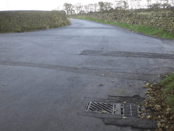 A rural road surface with a gully grating set into the channel, the tarmac around it patched and scarred, grass verge and hedge behind.