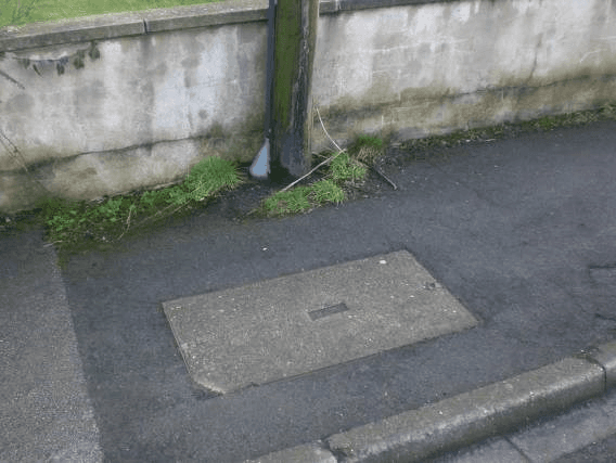 A footway against a rendered wall, with a rectangular inspection cover set flush in the paving and a service pipe running down the wall to it.
