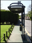 A bus shelter with an illuminated route sign, standing on a footpath with bollards and a clipped hedge alongside.