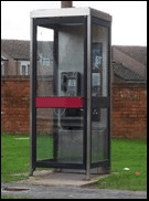 A red telephone kiosk standing on a grass verge in front of a brick wall.