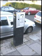 A pay and display parking machine on a paved parking area, cars parked alongside.